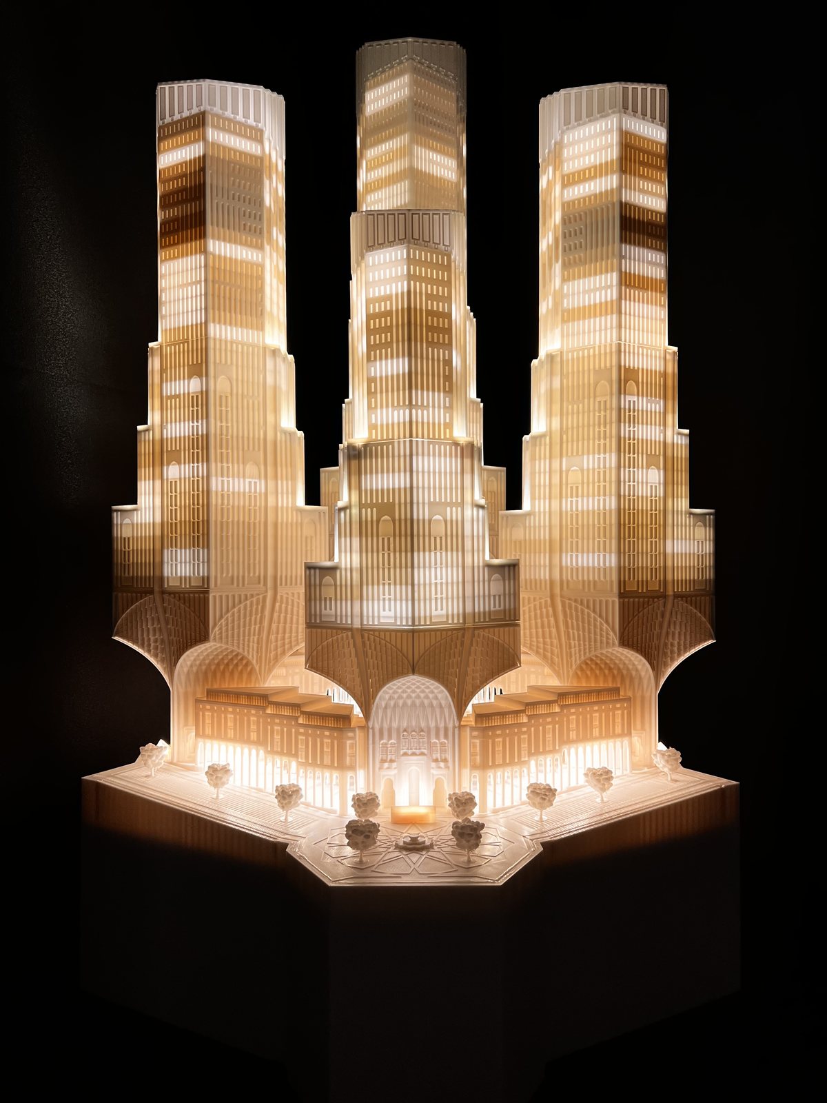 Illuminated physical scale model of three towers with arched bases and a landscaped plaza